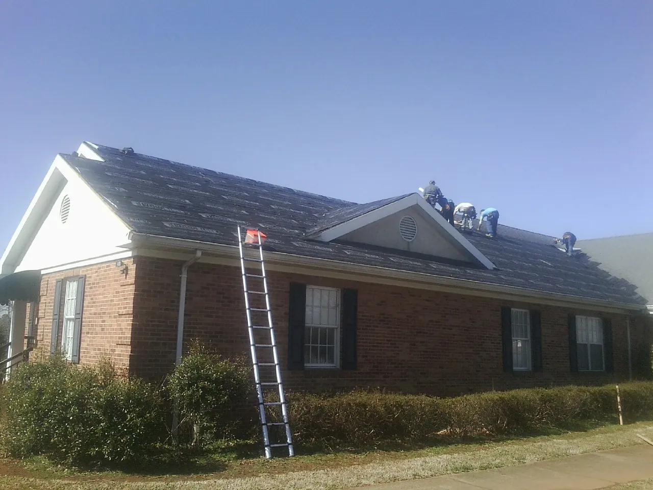 Crew tearing shingles off a brick ranch roof with a ladder set — Emergency Roof Repair work by our crew in Hubbardston