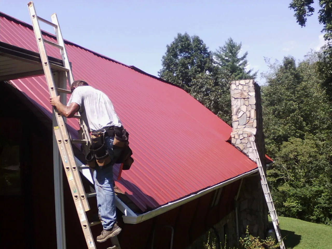Roofer on a ladder beside a red metal roof and stone chimney — Soffit & Fascia Repair by Roofing Brothers in Hubbardston